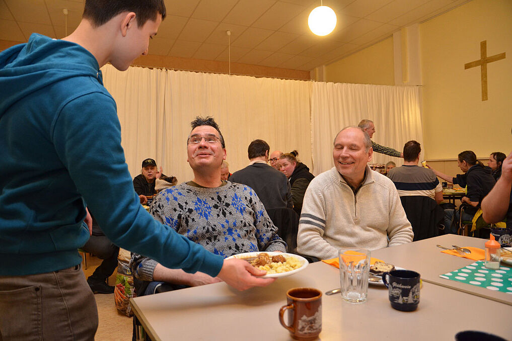 Colin bringt den Gästen ihr warmes Essen, während sich Pfarrer Kalle Lenz (r.) mit den Gästen ins Gespräch kommt. (Foto: Alfred Herrmann)