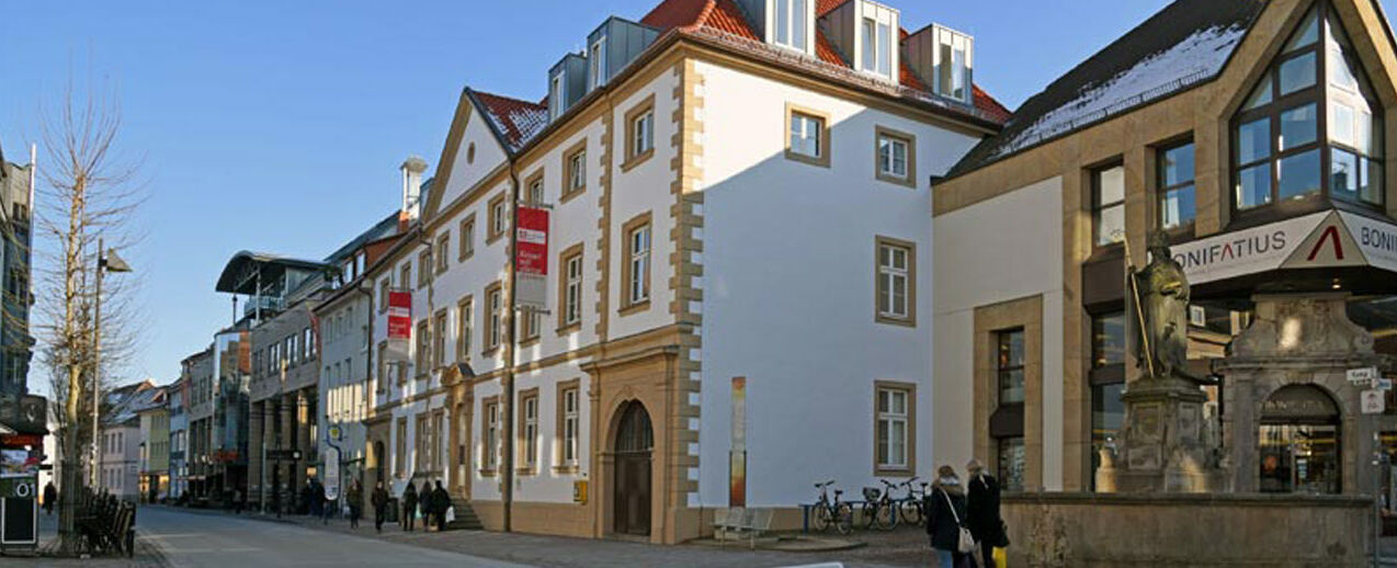 Das Gebäude am Kamp 22, das heutige Bonifatiushaus, wurde um 1720 erbaut. Die Tafel im Vordergrund zeigt eine Ansicht des durch Bomben zerstörten Gebäudes nach dem 2. Weltkrieg, das in den Jahren 1947-49 durch das Bonifatiuswerk wieder aufgebaut wurde. Foto: Patrick Kleibold