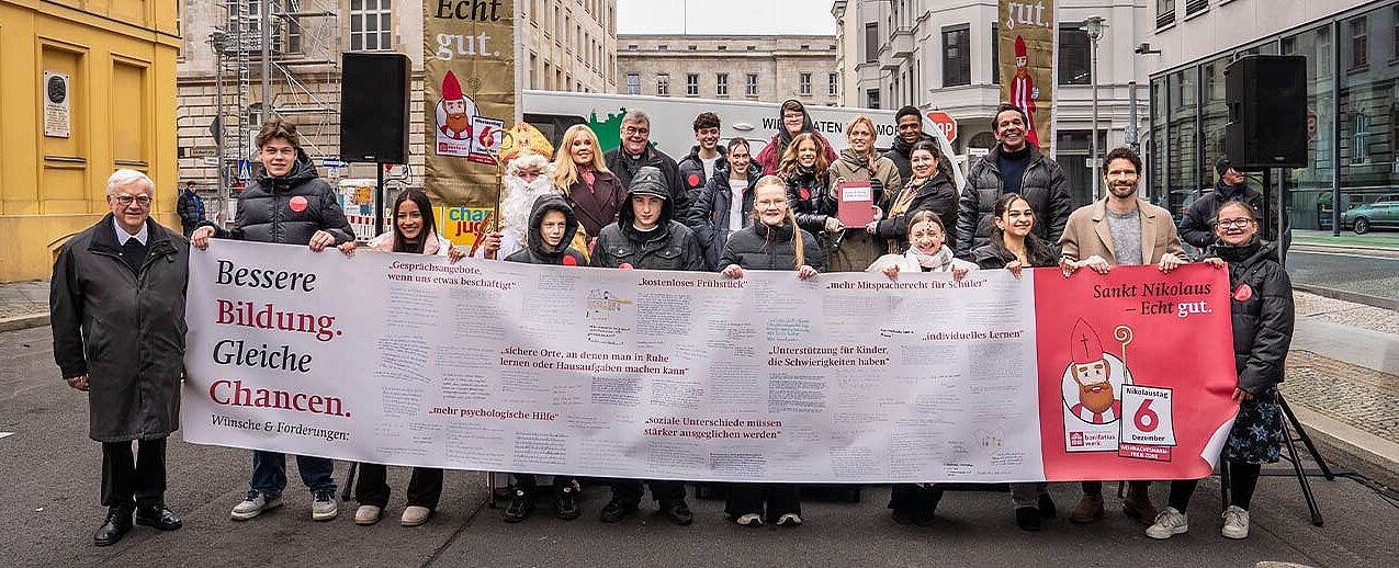 Höhepunkt der bundesweiten Nikolausaktion des Bonifatiuswerkes in Berlin: Auf einem fünf Meter langen Banner überreichten Jugendliche ihre Forderungen für bessere Bildung und gleiche Chancen an Bundesjugendministerin Karin Prien (letzte Reihe mit Buch). Mit im Bild: Maite Kelly (Botschafterin der Nikolausaktion, hintere Reihe 2.v.l.), Bonifatiuswerk-Generalsekretär Monsignore Georg Austen (hintere Reihe 3.v.l.), Erzbischof Dr. Heiner Koch (links), Ex-Fußballprofi und Stiftungsgründer Arne Friedrich (2. v.r.) und Moderator Juri Tetzlaff (3. v.r.). Foto: Hartmut Salzmann