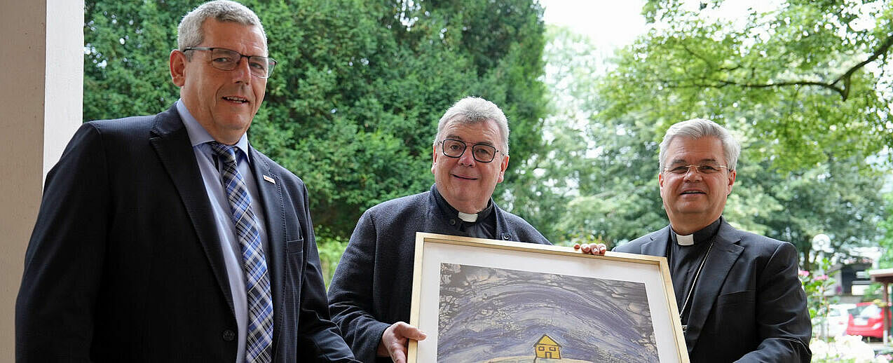 Bonifatiuswerk-Geschäftsführer Ingo Imenkämper (von links) und Bonifatiuswerk-Generalsekretär Monsignore Georg Austen übergeben das Lindenberg-Bild "Dach der Welt" an den Paderborner Erzbischof Dr. Udo Markus Bentz. (Foto: Matthias Band)