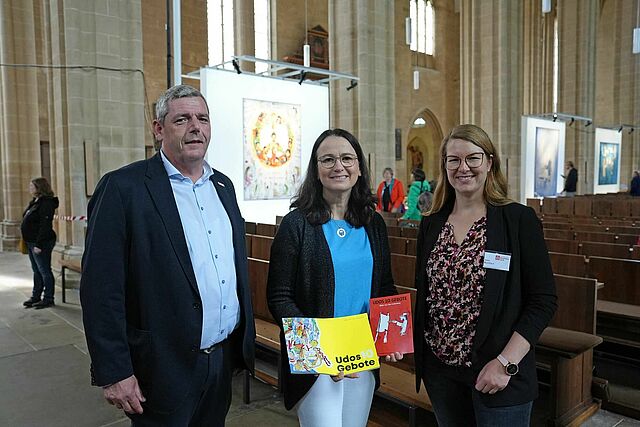 Ingo Imenkämper (links) und Kathrin Keller (rechts) überreichten der 12.500 Besucherin Christel Schmidt einen Katalog zur Ausstellung „Udos 10 Gebote“ und ein limitiertes Briefmarkenheft. (Foto: Simon Helmers)