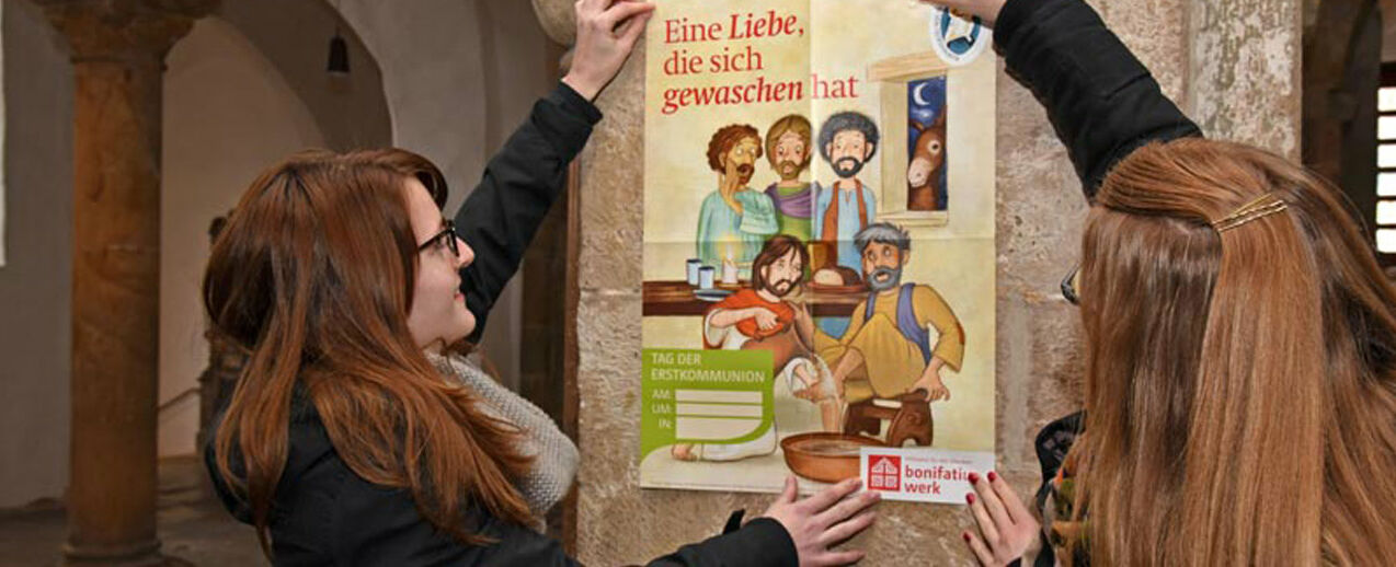 Ann-Kathrin Reker (Links) und Lena Reiher aus dem Bonifatiuswerk präsentieren das diesjährige Plakat zur Kommunionaktion, das die Geschichte der Fußwaschung aus dem Johannesevangelium zeigt. Foto: Patrick Kleibold 