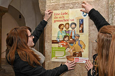 Ann-Kathrin Reker (Links) und Lena Reiher aus dem Bonifatiuswerk präsentieren das diesjährige Plakat zur Kommunionaktion, das die Geschichte der Fußwaschung aus dem Johannesevangelium zeigt. Foto: Patrick Kleibold 