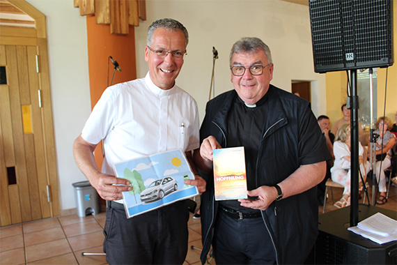 Monsignore Georg Austen überreicht Pater Christian Heim symbolisch ein Bild eines E-Autos, welches vom Bonifatiuswerk bewilligt wurde. (Foto: Oliver Gierens)